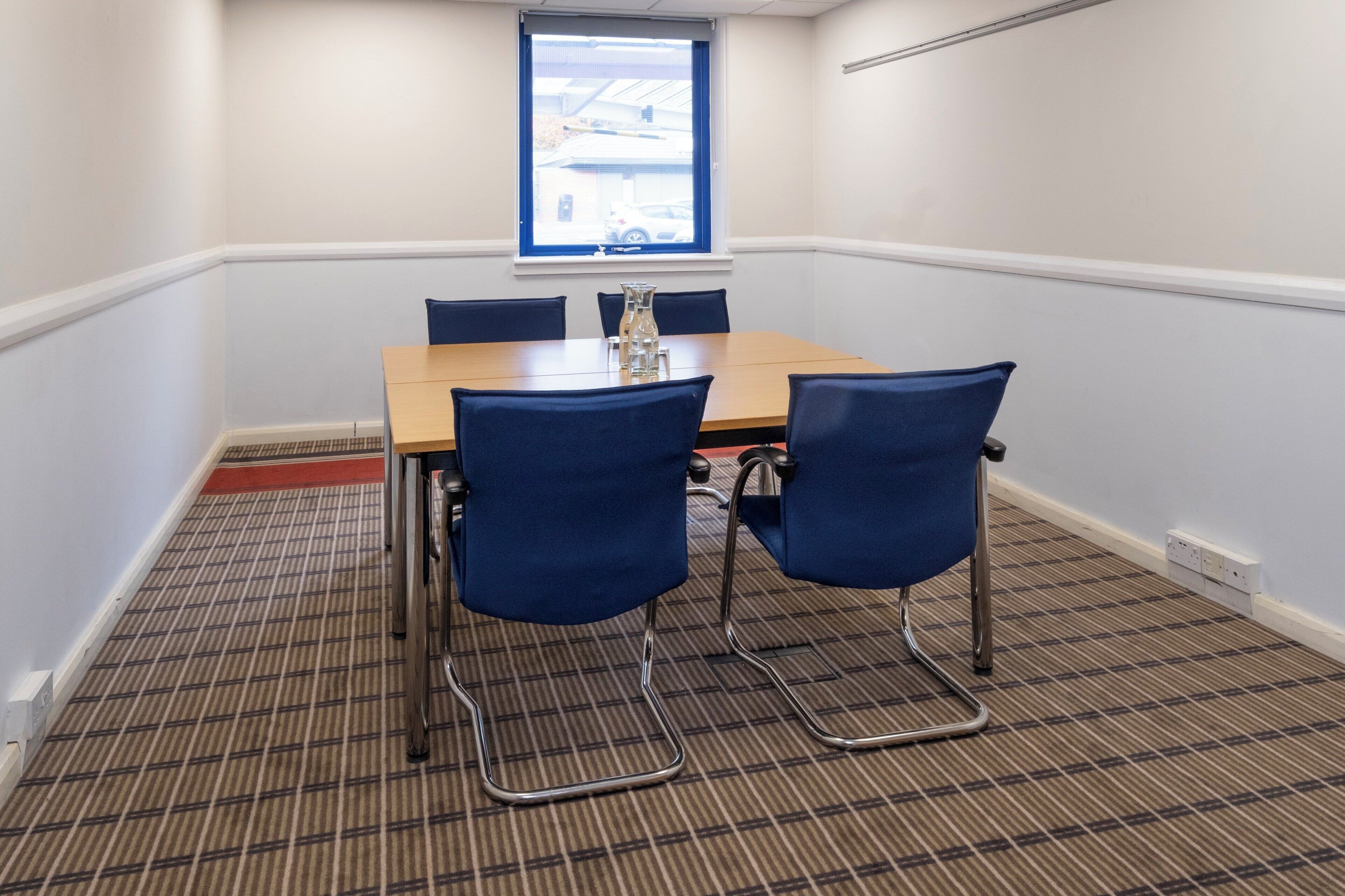 A conference room with a wooden table, four blue chairs, a window with a view, and a patterned carpet.