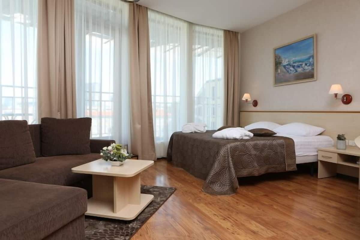 A hotel room with a sofa, a bed with a patterned bedspread, a small table with a vase, and a painting on the wall.
