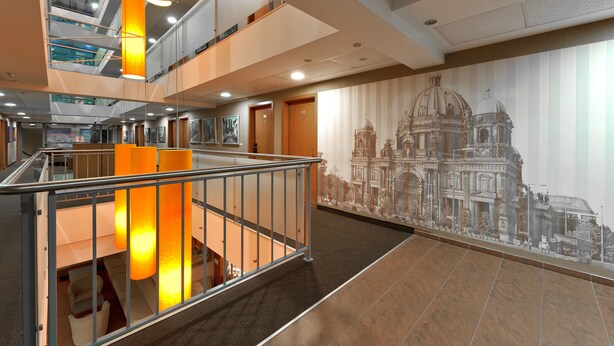 Un hall d’hôtel à plusieurs niveaux, doté d’un escalier, d’une fresque représentant un bâtiment imposant et d’un design moderne.