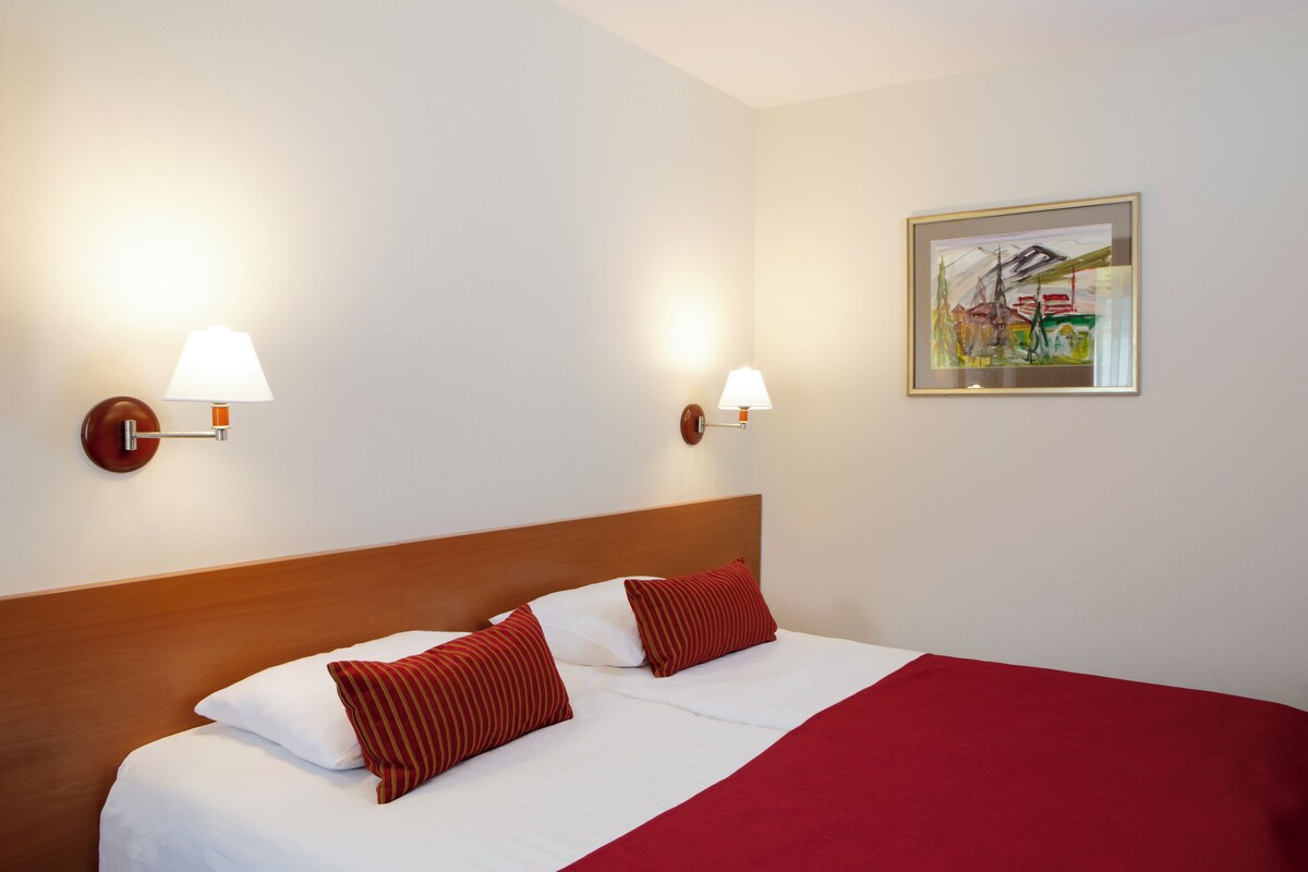Ein Hotelzimmer mit Holz-Kopfteil, weißer Bettwäsche, roten Kissen, zwei Wandlampen und einem gerahmten Bild an der Wand.