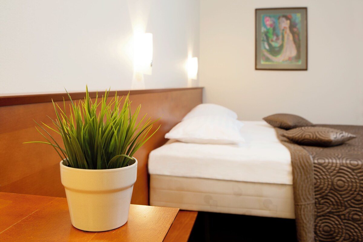 Ein ordentlich bezogenes Bett mit einem weißen Kopfkissen und einer braunen Decke, ein weißes Blumentopf auf einem Holztisch und ein Bild an der Wand.