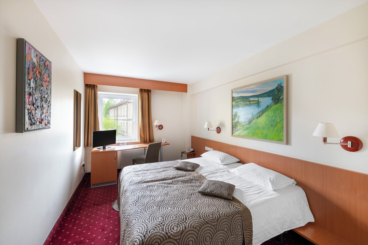 Quarto de hotel com cama, escrivaninha e um quadro na parede.