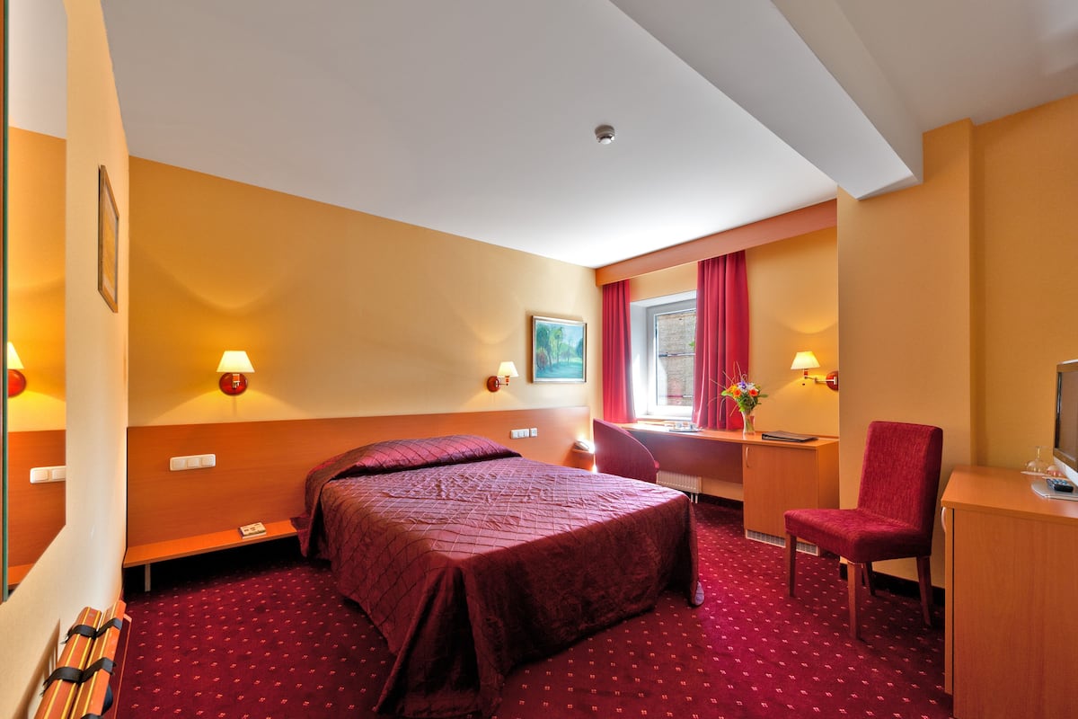 A hotel room with a large bed, a desk with a chair, a window with red curtains, and wall-mounted lamps.