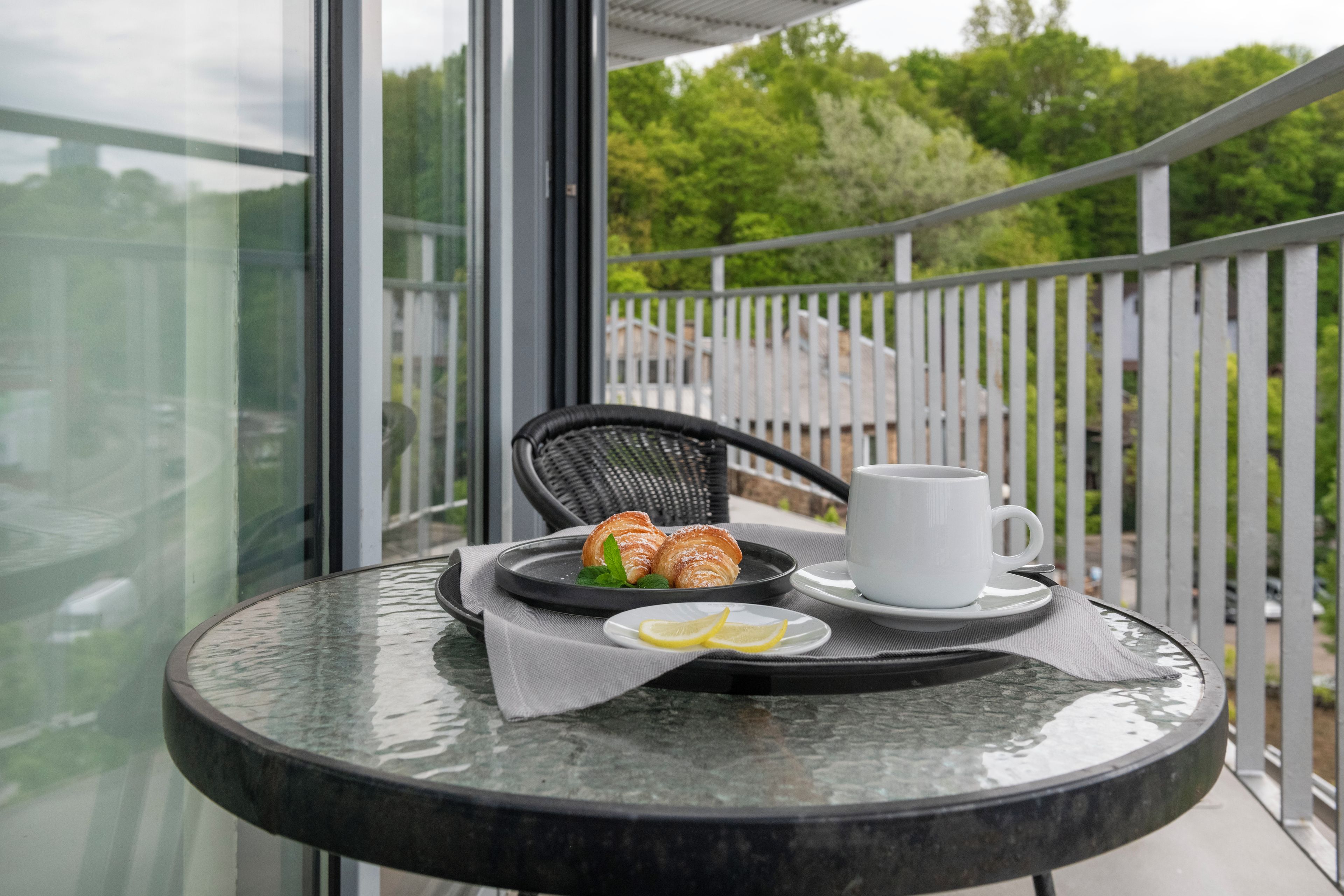 Ein runder Tisch mit einer weißen Tasse, einem Teller mit Croissants und einem Stück Zitrone, sowie ein Korbstuhl.