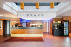 A hotel reception area with a reception desk, a clock displaying time in multiple cities, a vending machine, and a digital screen. - Art City Inn (Vilnius)