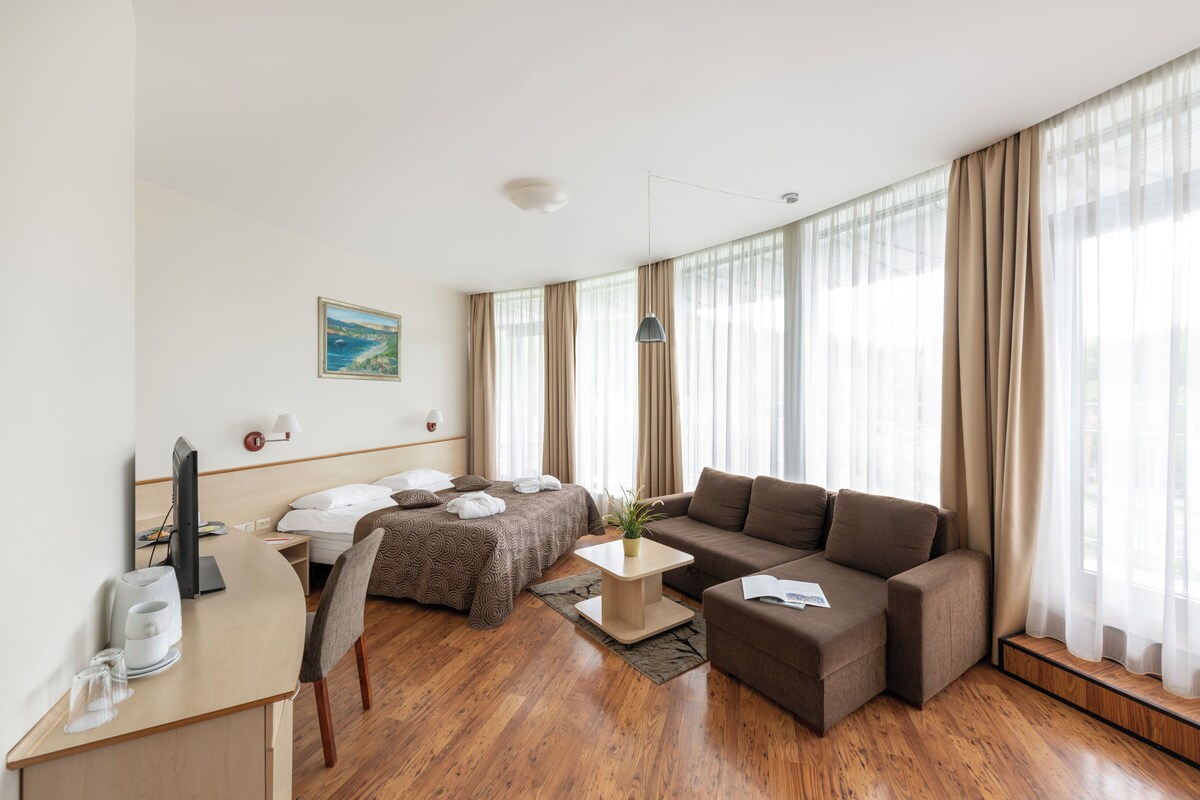 Quarto de hotel com cama, sofá, escrivaninha com luminária, cadeira e janela com cortinas.