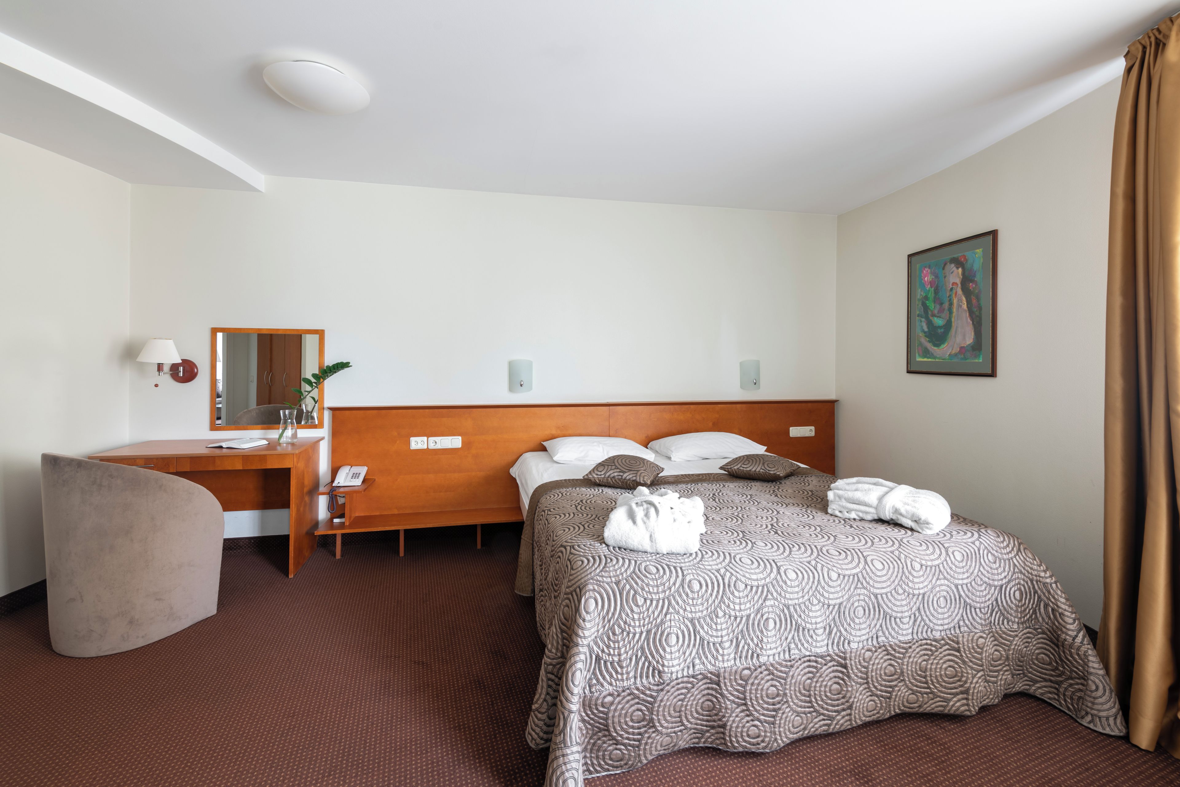 A hotel room with a bed, a desk, a chair, a mirror, and a painting on the wall.