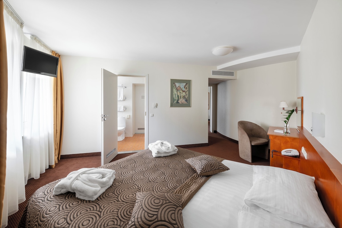 Quarto de hotel com cama, escrivaninha, cadeira, um quadro e banheiro visível através de uma porta aberta.