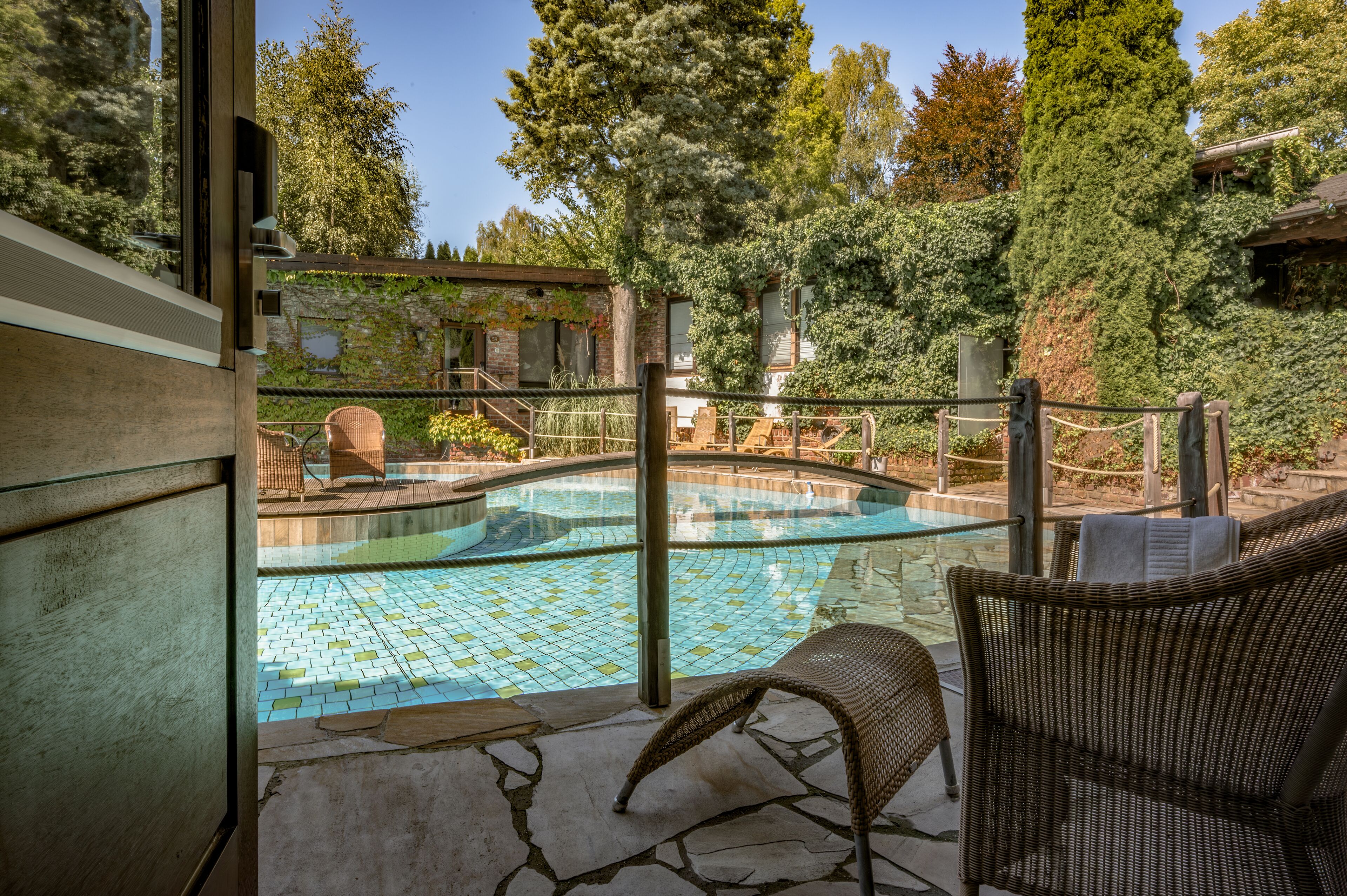 Una piscina exterior con solárium alicatado, mobiliario de mimbre y vistas a un edificio con paredes cubiertas de hiedra.