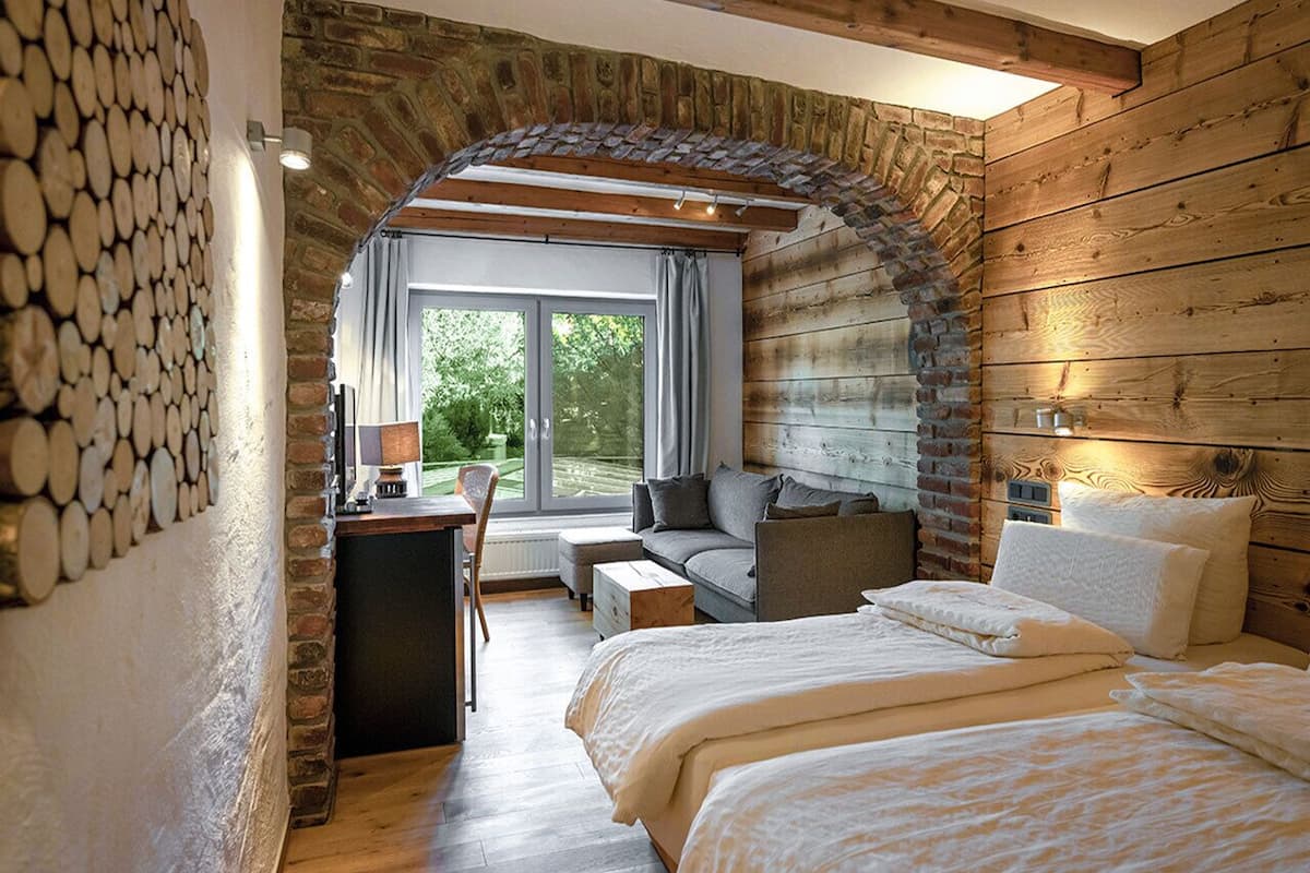 A cozy bedroom with a stone archway, a bed with white bedding, a wooden desk, and a view of greenery outside.