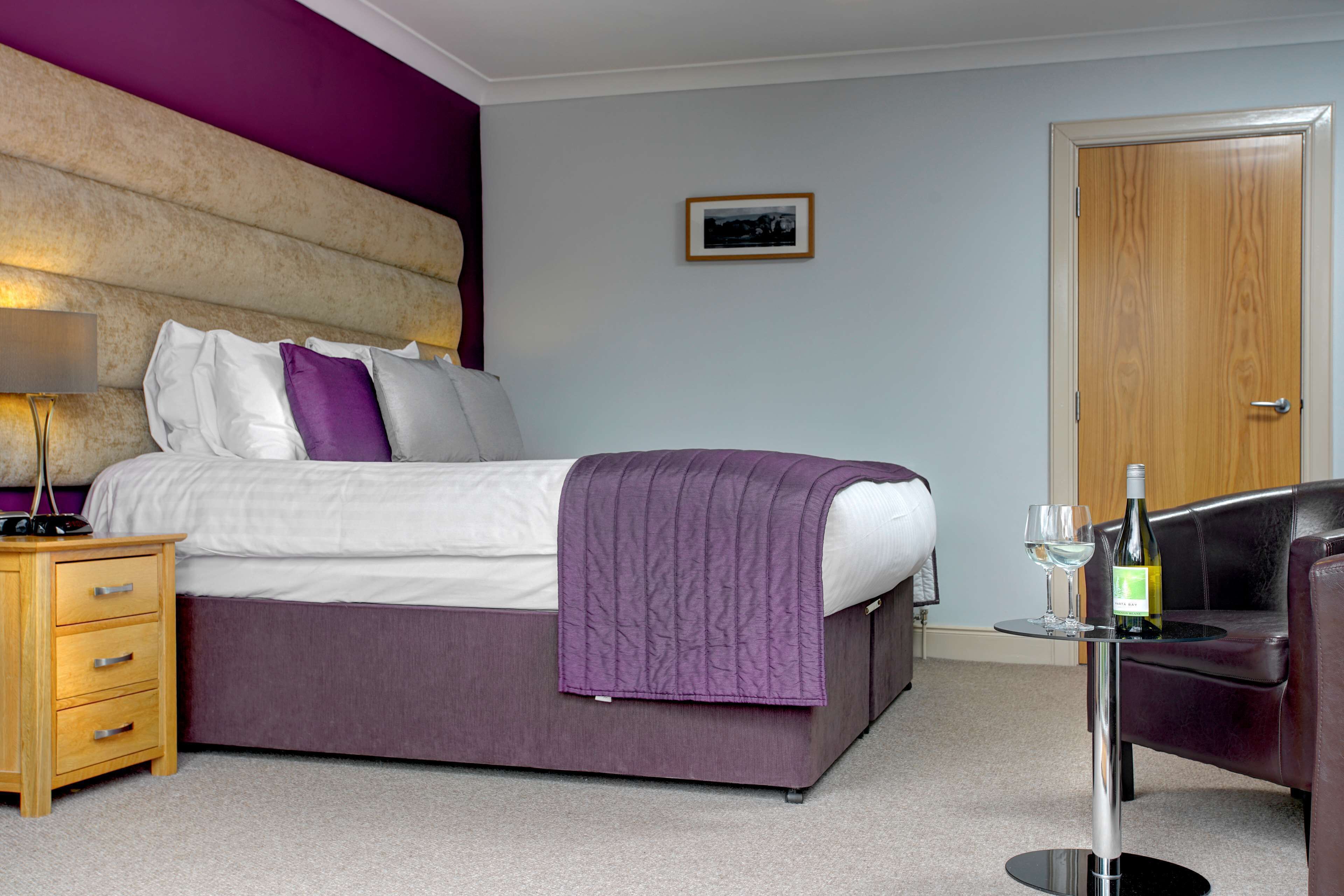 A hotel room with a bed, a nightstand, a table with a bottle and glass, and a door.