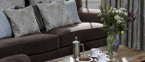 A cozy living room with a brown sofa, patterned cushions, a wooden coffee table with a magazine, a coffee pot, cups, a plate with cookies, and a vase with flowers.