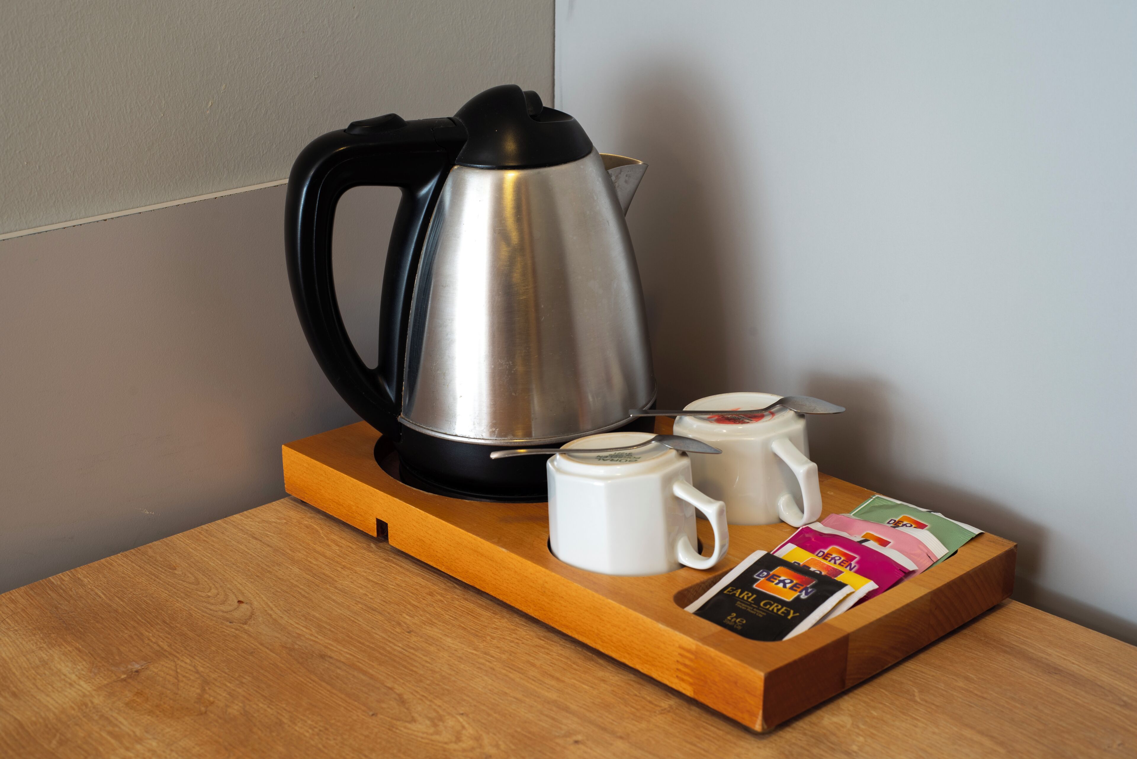 A stainless steel kettle on a wooden tray with two white mugs and a packet of coffee creamer.