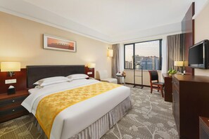 A hotel room with a large bed, a desk, a chair, a television, and a window with a city view.