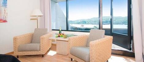 A room with two wicker armchairs, a small table with fruits, and a view of a lake through large windows.