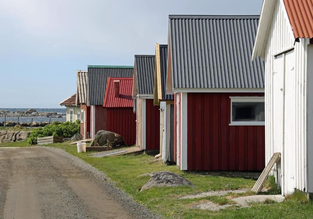 A row of red and white buildings with corrugated metal roofs, a gravel path, and a view of the sea in the background.