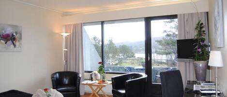 A hotel room with a bed, two black chairs, a small table with a vase, a desk with a lamp, a TV, and a large window with a view of trees and mountains.