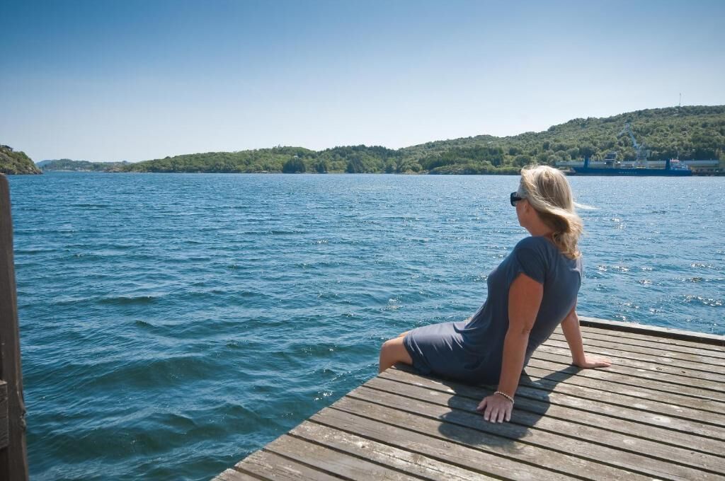 Een vrouw zittend op een houten steiger met uitzicht op een wateroppervlak en een heuvelachtig landschap op de achtergrond.