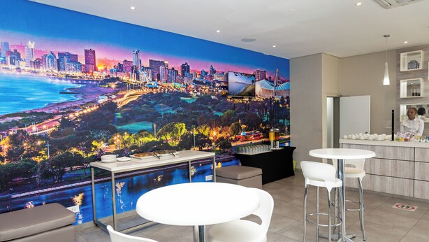 A modern hotel room with a large cityscape mural, a white dining area, and a kitchen counter. - The Riverside Hotel (Durban North)