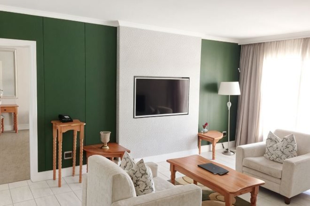 A hotel room with a green accent wall, a flat-screen TV, a sofa, a coffee table, a lamp, and a window with curtains.