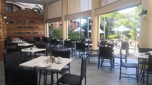 An indoor dining area with brick walls, large windows, and outdoor seating with umbrellas. Tables are set with white tablecloths and black chairs. - The Riverside Hotel (Durban North)