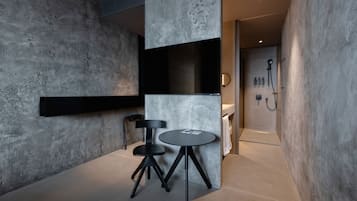 Un baño moderno con pared de concreto, un tocador negro, una mesa redonda con una silla y una zona de ducha con cabezal de ducha de mano.