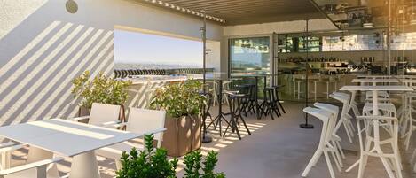 A rooftop bar with white tables and chairs, potted plants, and a view of the cityscape.