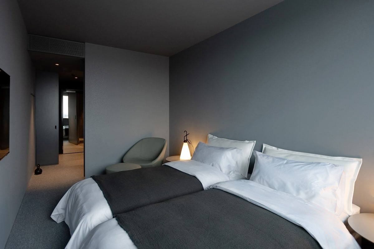 Una habitación de hotel con una cama grande, una silla, una mesita de noche y una lámpara.