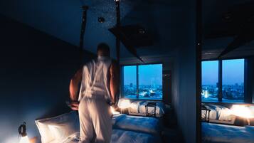 A modern hotel room with a city view, a large bed, a bedside table with a lamp, and a mirror reflecting the room.