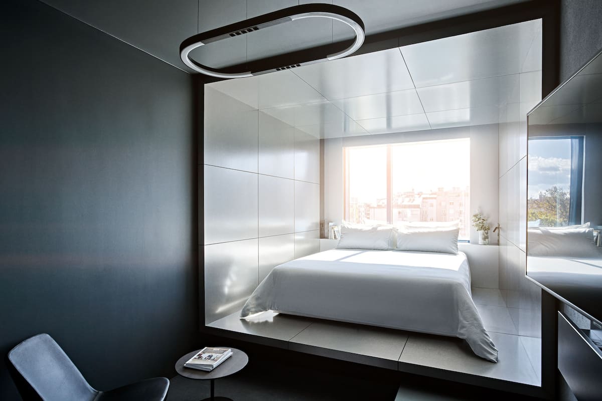 Una habitación de hotel moderna con una cama grande, una pequeña mesa redonda, una silla, un ventanal con vistas a la ciudad y un escritorio empotrado con espejo.