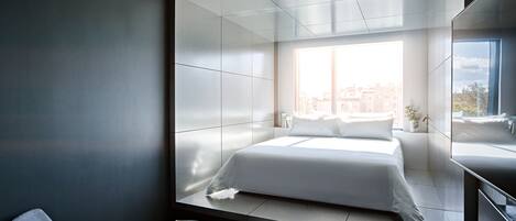 A modern hotel room with a large bed, a small round table, a chair, a large window with a city view, and a built-in desk with a mirror.