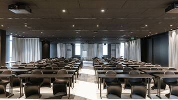 Une salle de conférence avec des rangées de tables et de chaises, de grandes fenêtres avec des rideaux et un projecteur au plafond.