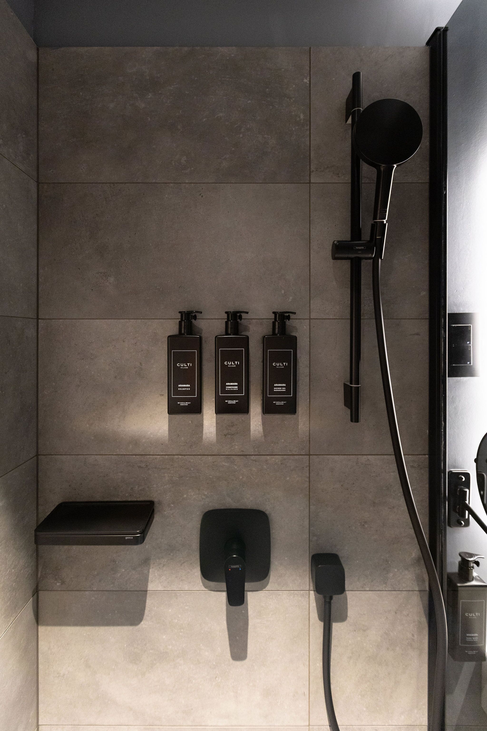 A modern bathroom with a sleek design, featuring a wall-mounted shower, a handheld showerhead, and three bottles of Culti shampoo.