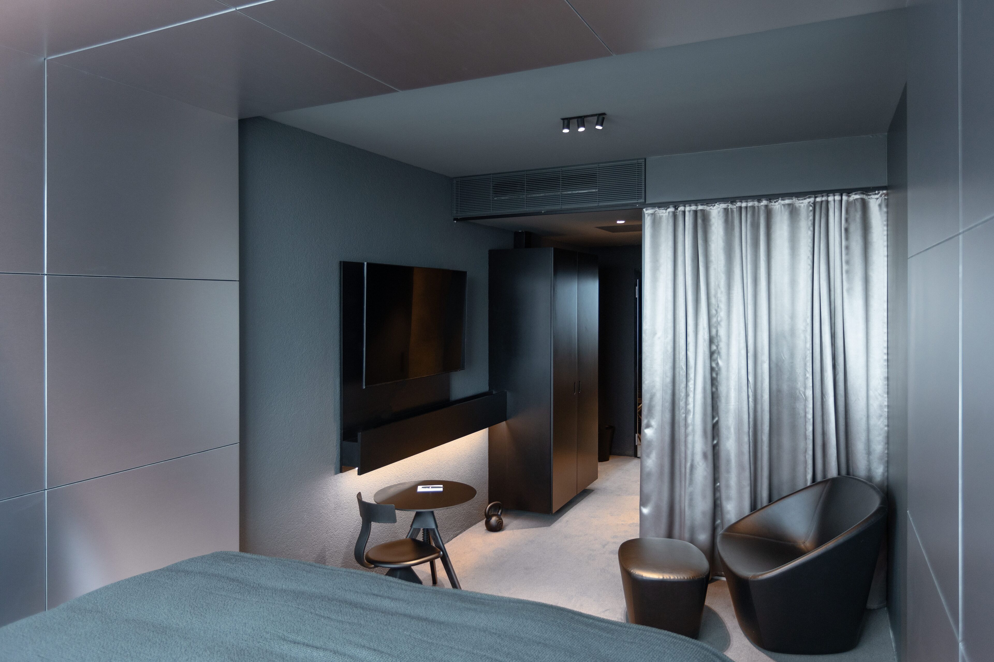 A modern hotel room with a bed, a TV mounted on the wall, a small round table with two chairs, and a large window with curtains.