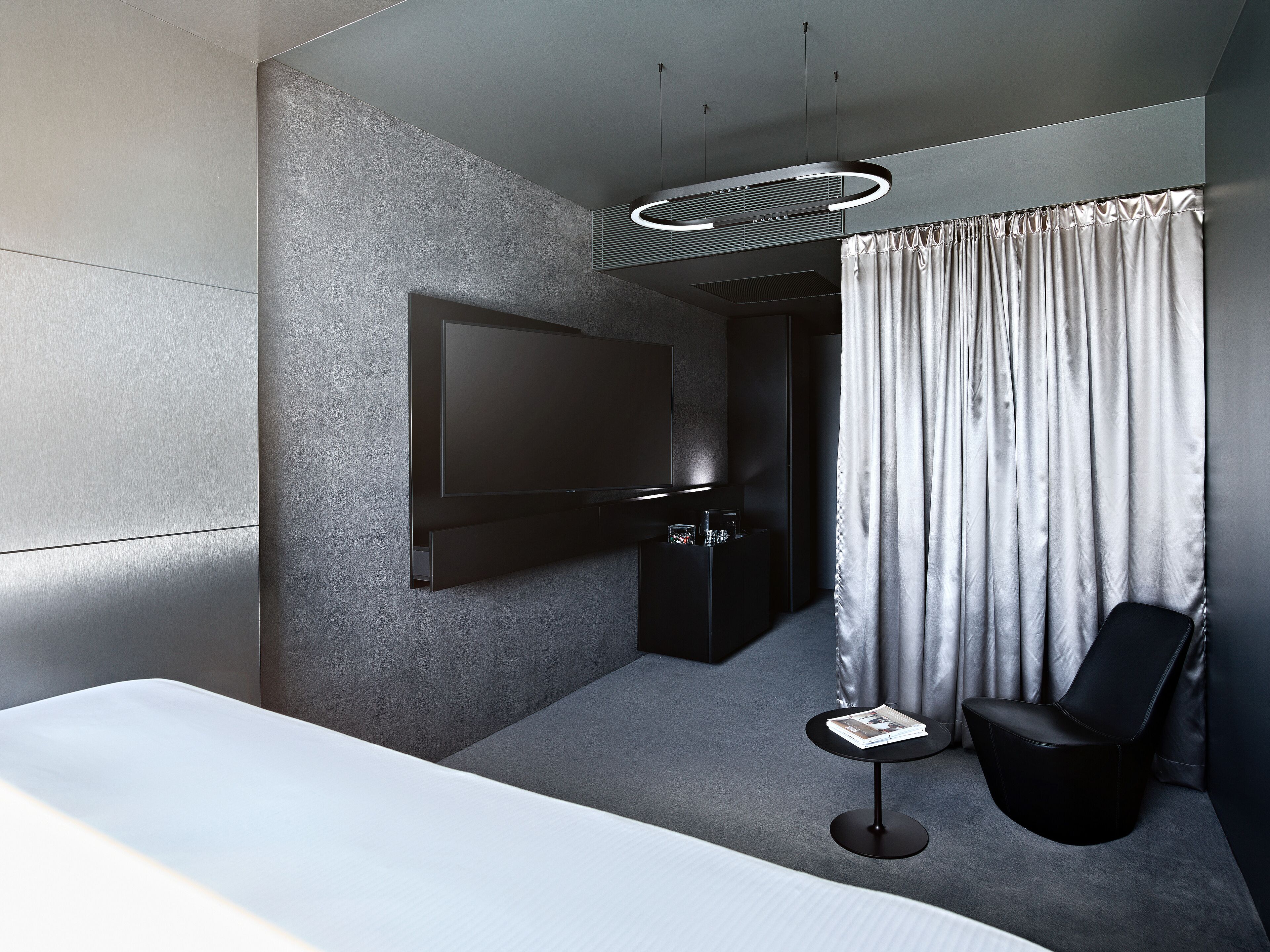 Una habitación de hotel moderna con una cama grande, un televisor de pantalla plana montado en la pared, una mesita de noche con una revista y un sillón de cuero negro.