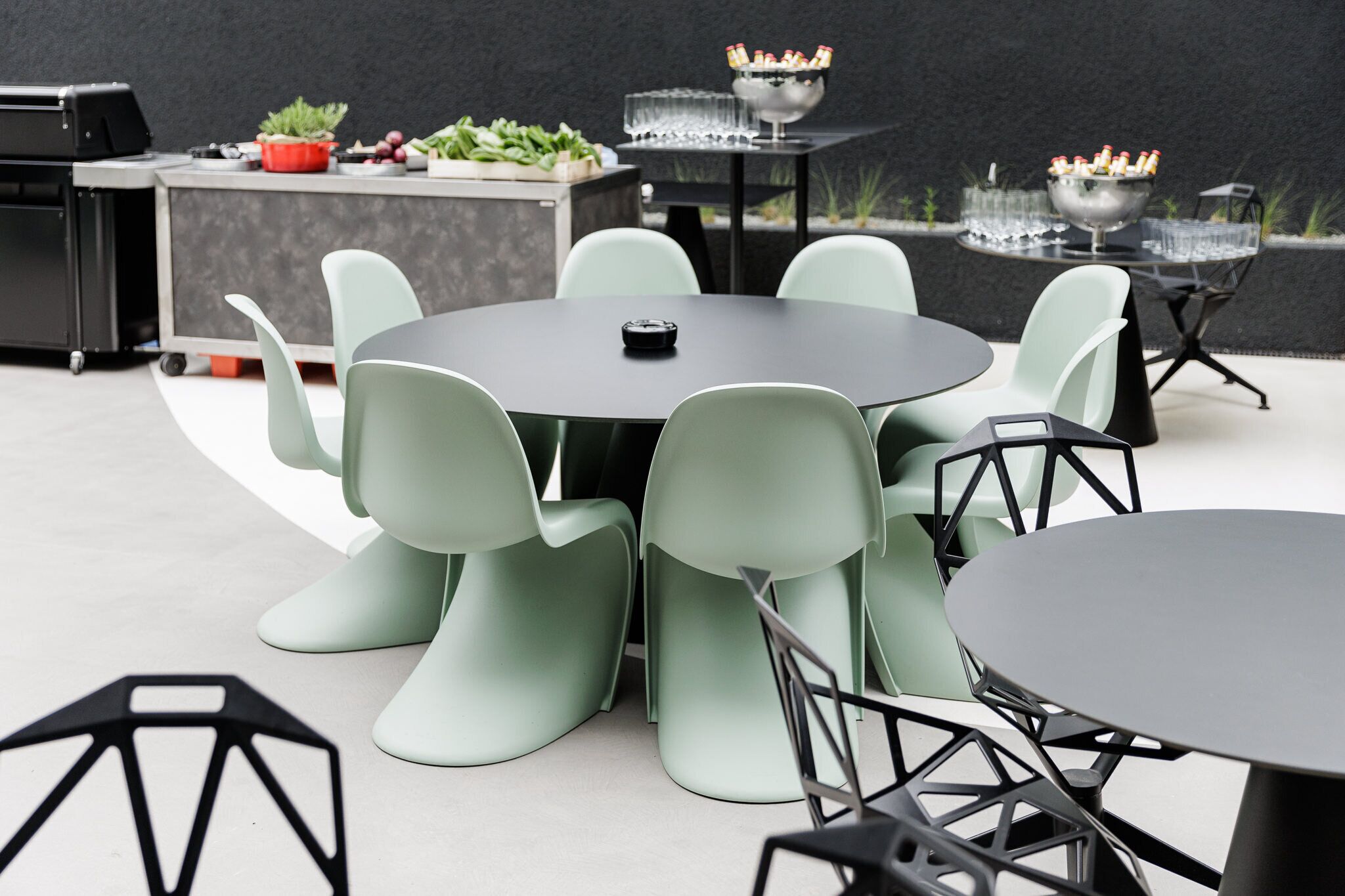 A modern outdoor dining area with round tables and light green chairs, surrounded by a black wall and a small garden.