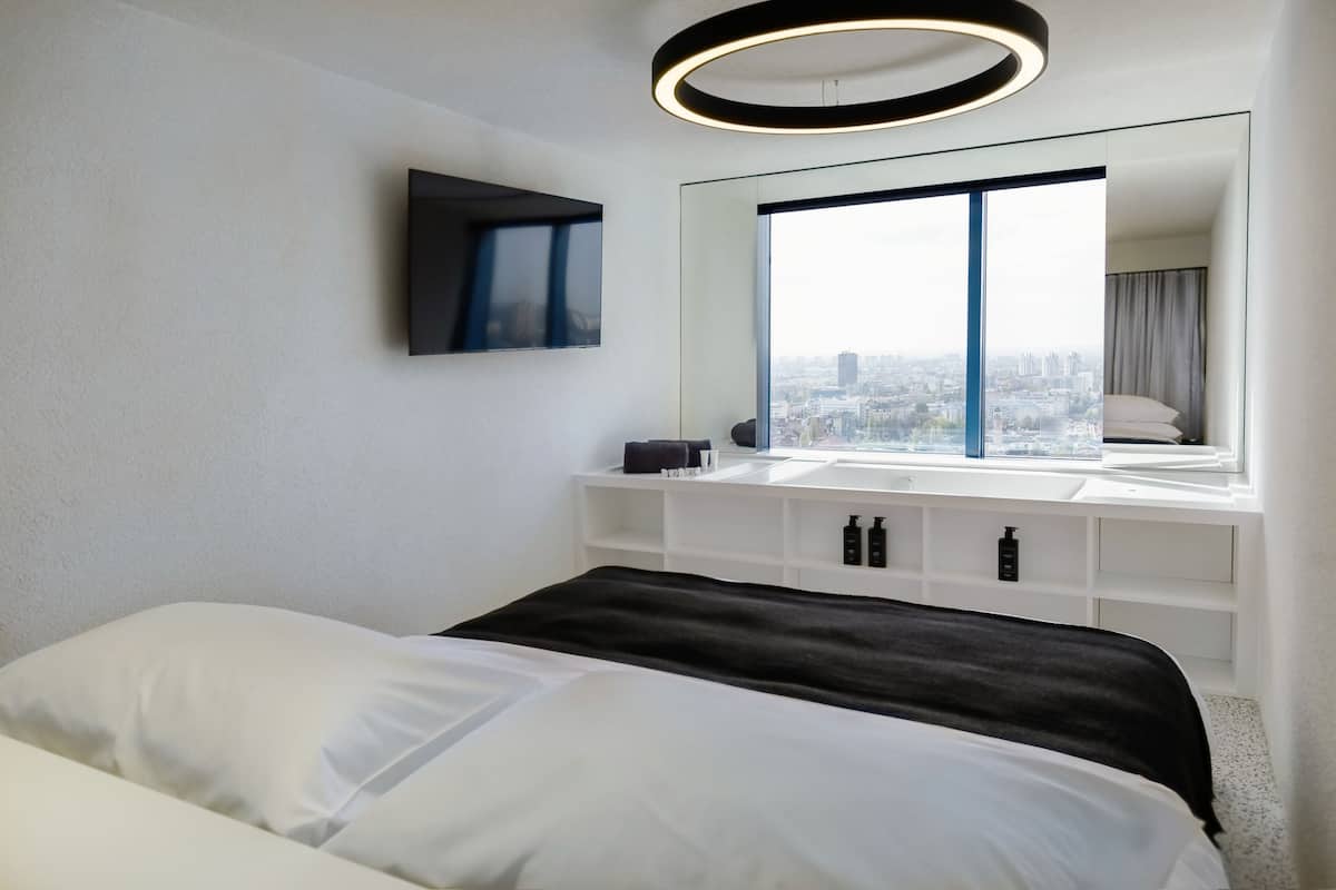 Una habitación de hotel moderna con una cama grande, vistas a la ciudad, una lámpara redonda y un espejo amplio.