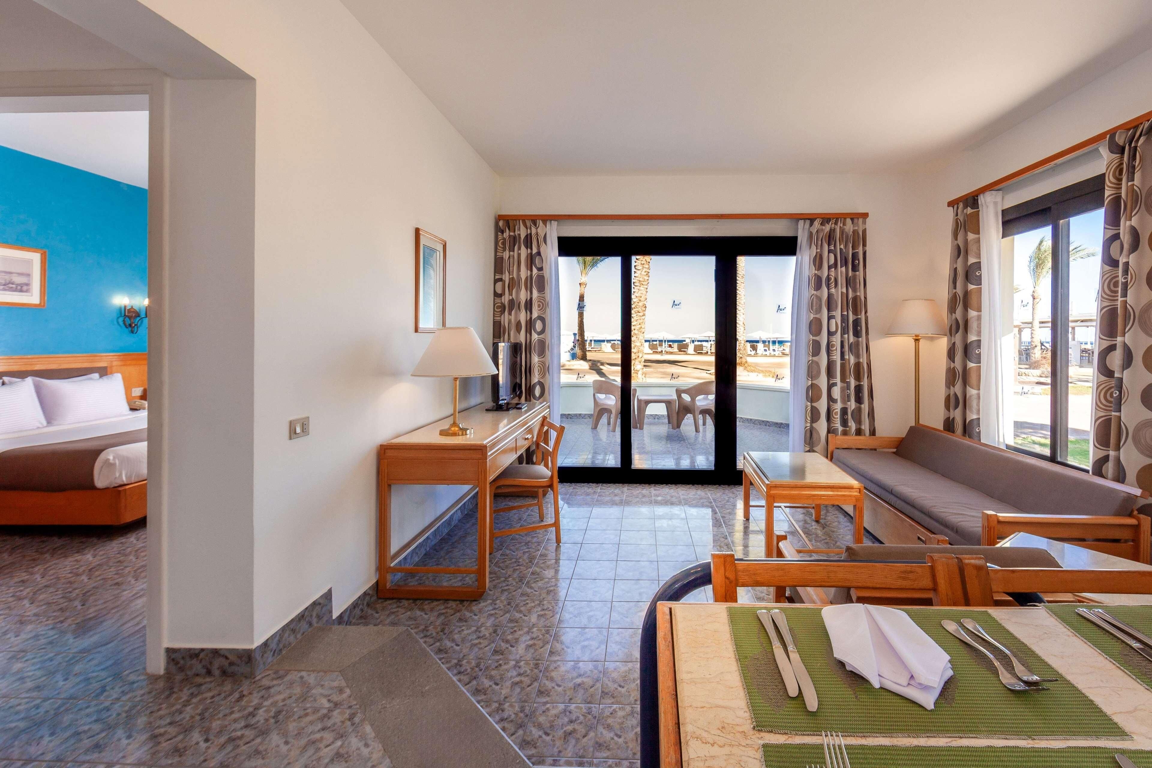 A hotel room with a bed, a desk, a chair, a sofa, and a dining area with a table set for two.