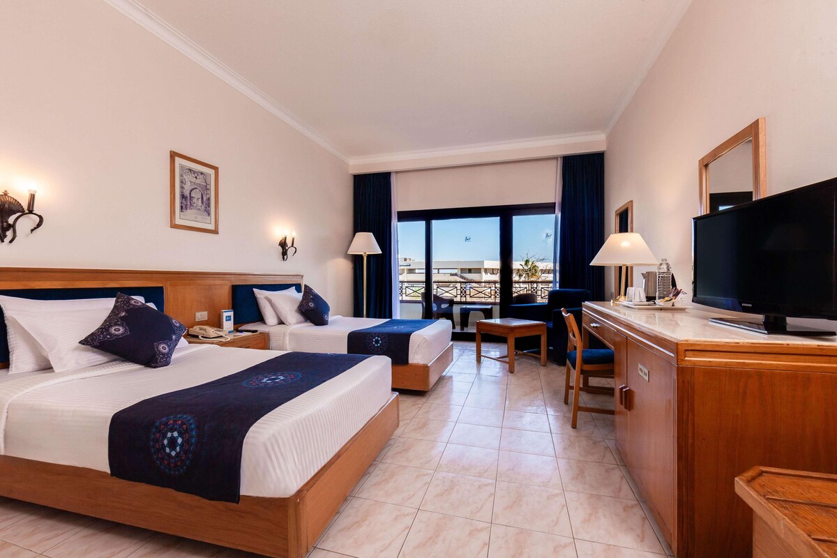 A hotel room with two beds, a desk, a television, and a balcony with a view.