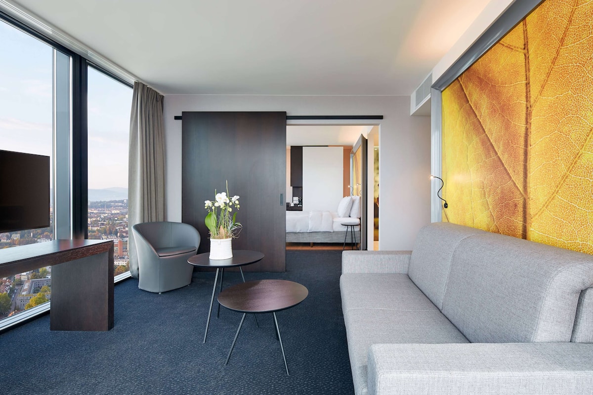 A modern hotel room with a large window, a grey sofa, a small table with a potted plant, and a view of the city.