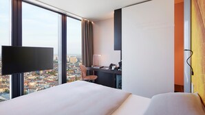 A hotel room with a large bed, a desk, a chair, a flat-screen TV, and a city view.