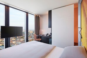 A hotel room with a large bed, a desk, a chair, a flat-screen TV, and a city view.