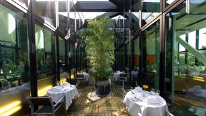An indoor dining area with tables set for two, a central potted plant, and large windows allowing natural light. - HYPERION Hotel Basel (Basel)