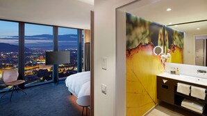 A hotel room with a large window offering a city view at night, a bed, a chair, a bathroom with a mirror and sink, and a wall-mounted TV. - HYPERION Hotel Basel (Basel)