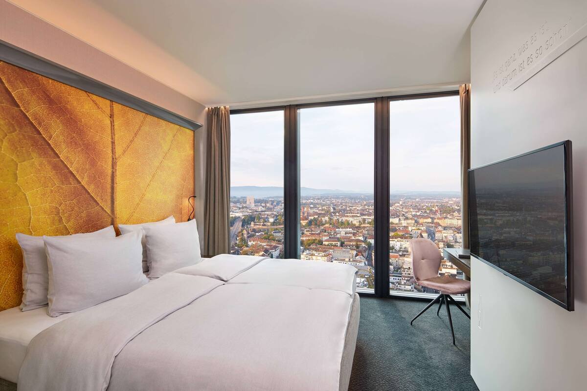 A hotel room with a large bed, a desk, and a chair. There is a large window with a city view.
