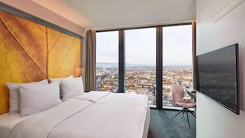 Une chambre d’hôtel avec un grand lit, un bureau et une chaise. Une grande fenêtre offre une vue sur la ville.