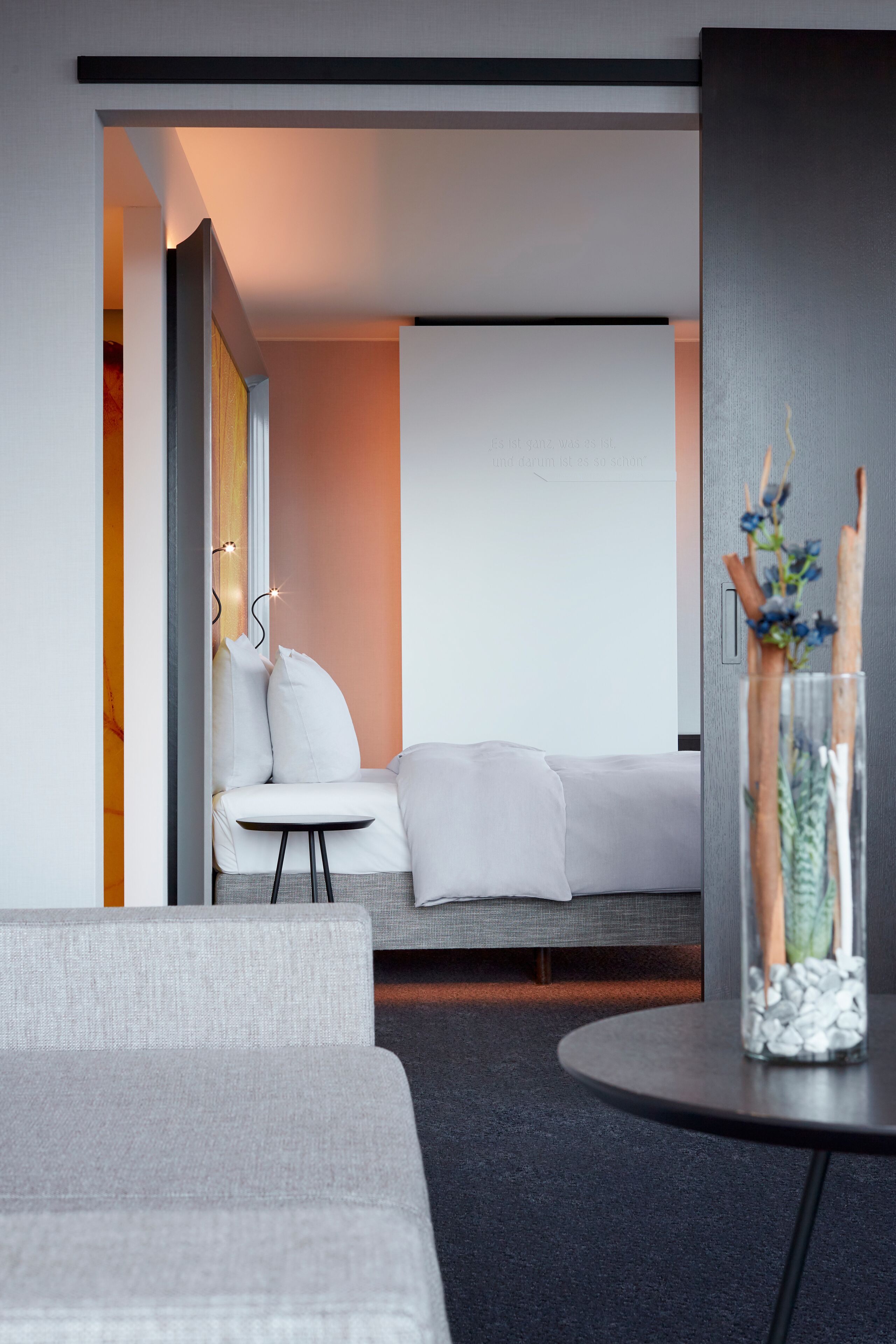 A modern hotel room with a grey sofa, a small round table with a vase, and a bed with white bedding.