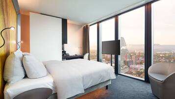 Une chambre d’hôtel moderne dotée d’un grand lit, d’un bureau, d’une chaise et offrant une vue sur le paysage urbain grâce à de grandes fenêtres.