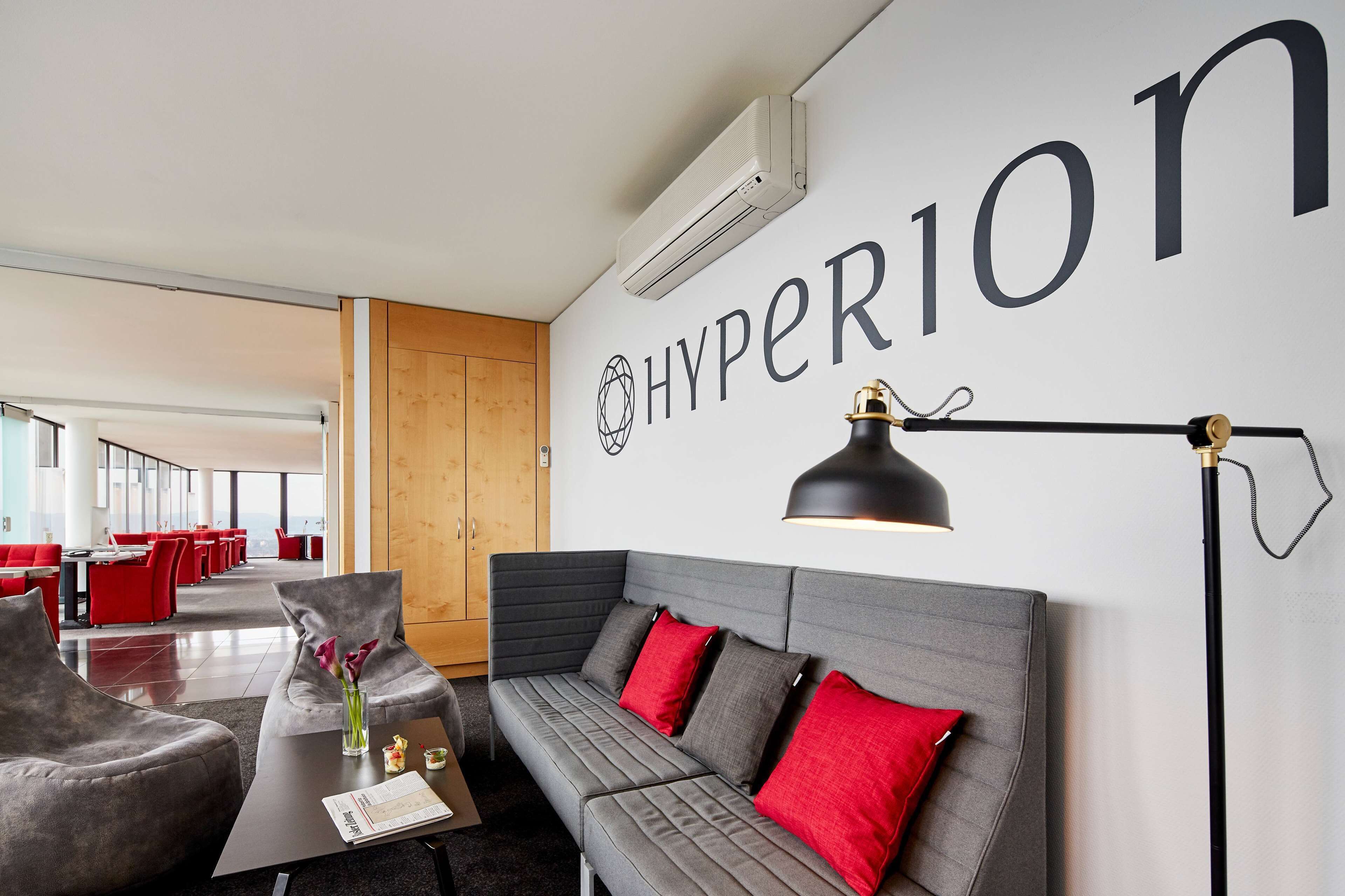 A modern hotel lobby with a grey sofa, red cushions, a black floor lamp, and a large wall sign reading "HYPERION".