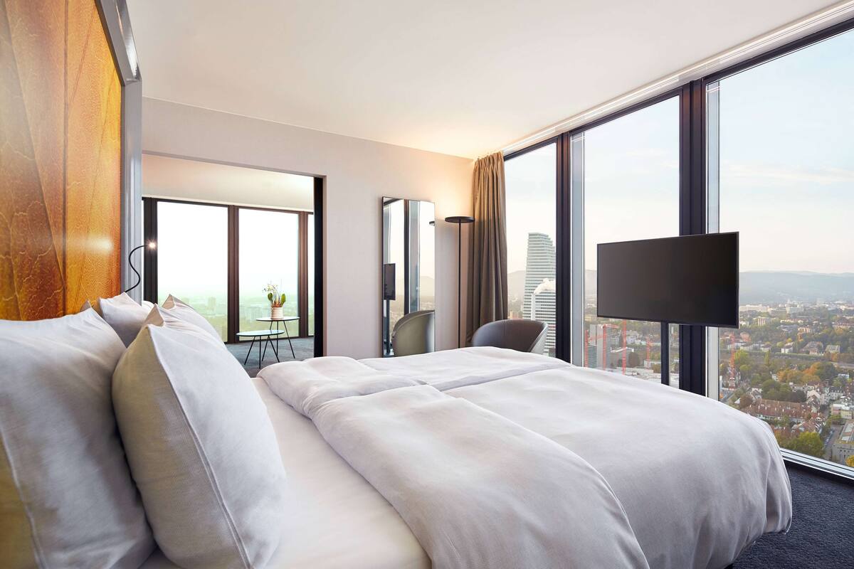 A hotel room with a large bed, a TV, a desk, and a view of the city.